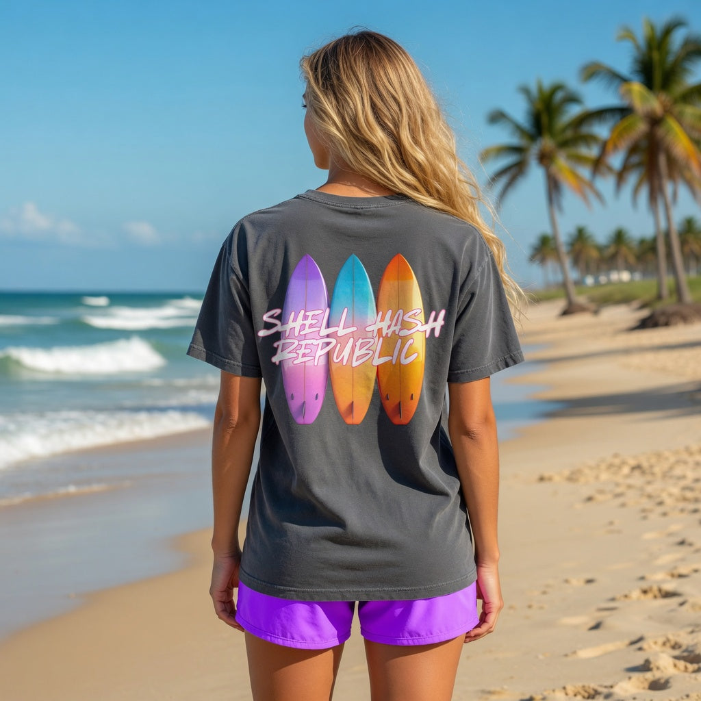 Woman in a black t-shirt with three surfboards and Shell Hash Republic text on the back, standing on a beach with palm trees