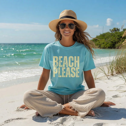 Woman wearing a Shell Hash Republic Comfort Colors T-Shirt with Beach Please graphic, straw hat, and sunglasses, sitting cross-legged on a sandy beach with the ocean in the background