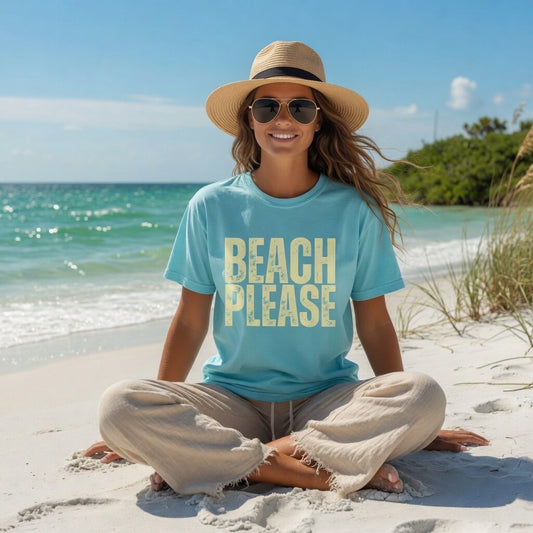Woman wearing a Shell Hash Republic Comfort Colors T-Shirt with Beach Please graphic, straw hat, and sunglasses, sitting cross-legged on a sandy beach with the ocean in the background