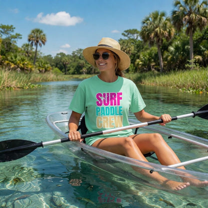 Woman in a Shell Hash Republic Comfort Colors Neon Surf Paddle Crew T-Shirt and straw hat, paddling a clear kayak on a clear river with palm trees in the background