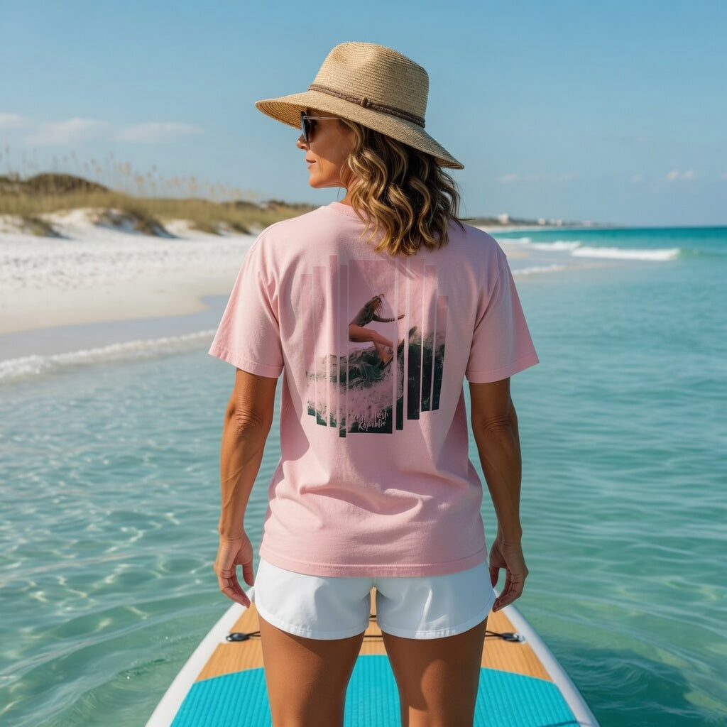 Woman in Shell Hash Republic Pink Wave Comfort Colors T-Shirt with Surfer Girl on Big Wave graphic, standing on a paddleboard in clear ocean water