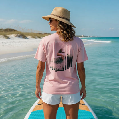 Woman in Shell Hash Republic Pink Wave Comfort Colors T-Shirt with Surfer Girl on Big Wave graphic, standing on a paddleboard in clear ocean water