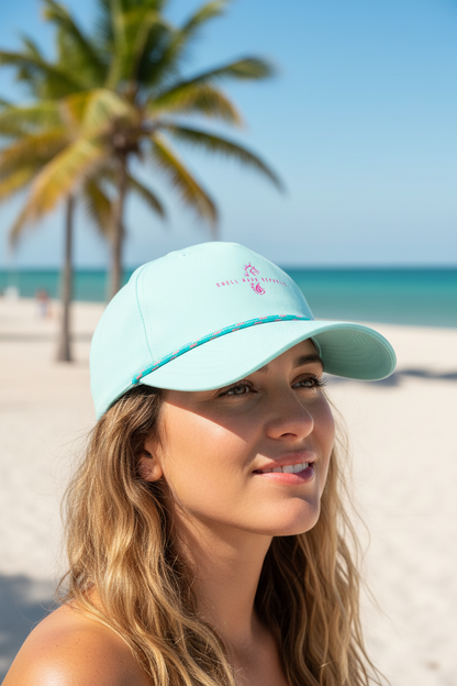 Woman wearing a Shell Hash Republic Small Fit Women's Hat by Imperial, light blue with pink logo, on a beach with palm trees and ocean in the background