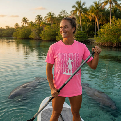 Woman on paddleboard with manatees, wearing Shell Hash Republic Neon Comfort Colors T-Shirt with Forever Chasing Sunsets graphic