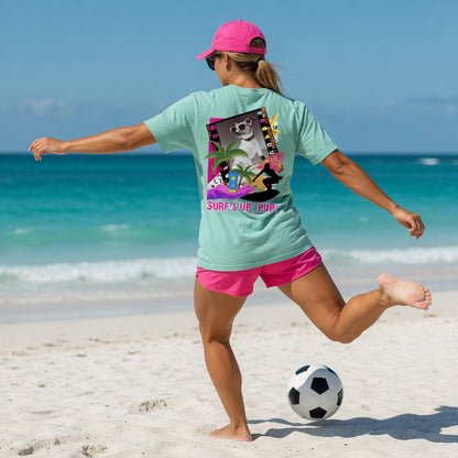 Woman in Shell Hash Republic Comfort Colors T-Shirt with Surfs Up Pup graphic, pink shorts, and pink cap, kicking a soccer ball on a sandy beach