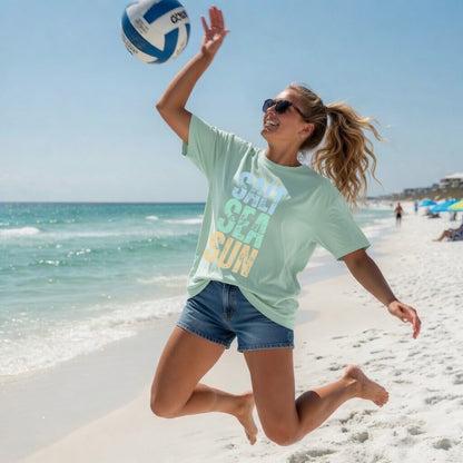 Woman jumping on a beach, wearing a Shell Hash Republic Neon Comfort Colors T-Shirt with a Salt, Sea, Sun graphic, hitting a volleyball