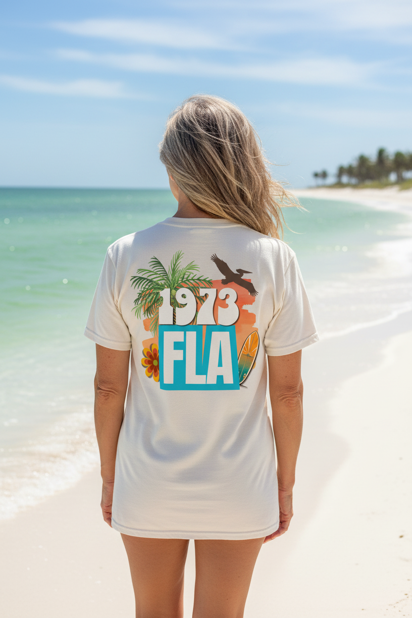 Woman from behind wearing a Shell Hash Republic Ivory Comfort Colors T-Shirt with a 1970s retro graphic of 1973 FLA, a palm tree, pelican, flower, and surfboard on a beach