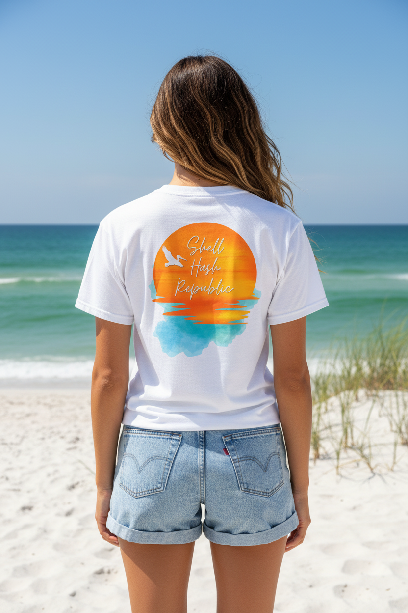 Woman from behind wearing a Shell Hash Republic White Comfort Colors T-Shirt with a Pelican Sunset graphic, standing on a beach