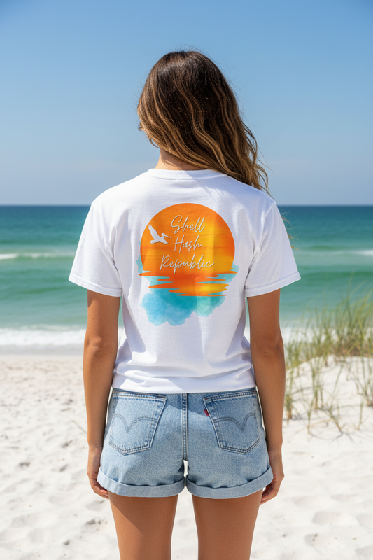Woman from behind wearing a Shell Hash Republic White Comfort Colors T-Shirt with a Pelican Sunset graphic, standing on a beach