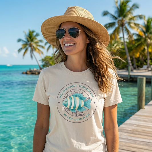 Woman wearing Shell Hash Republic Comfort Colors Ivory T-Shirt with striped reef fish graphic, standing on a dock by turquoise water and palm trees